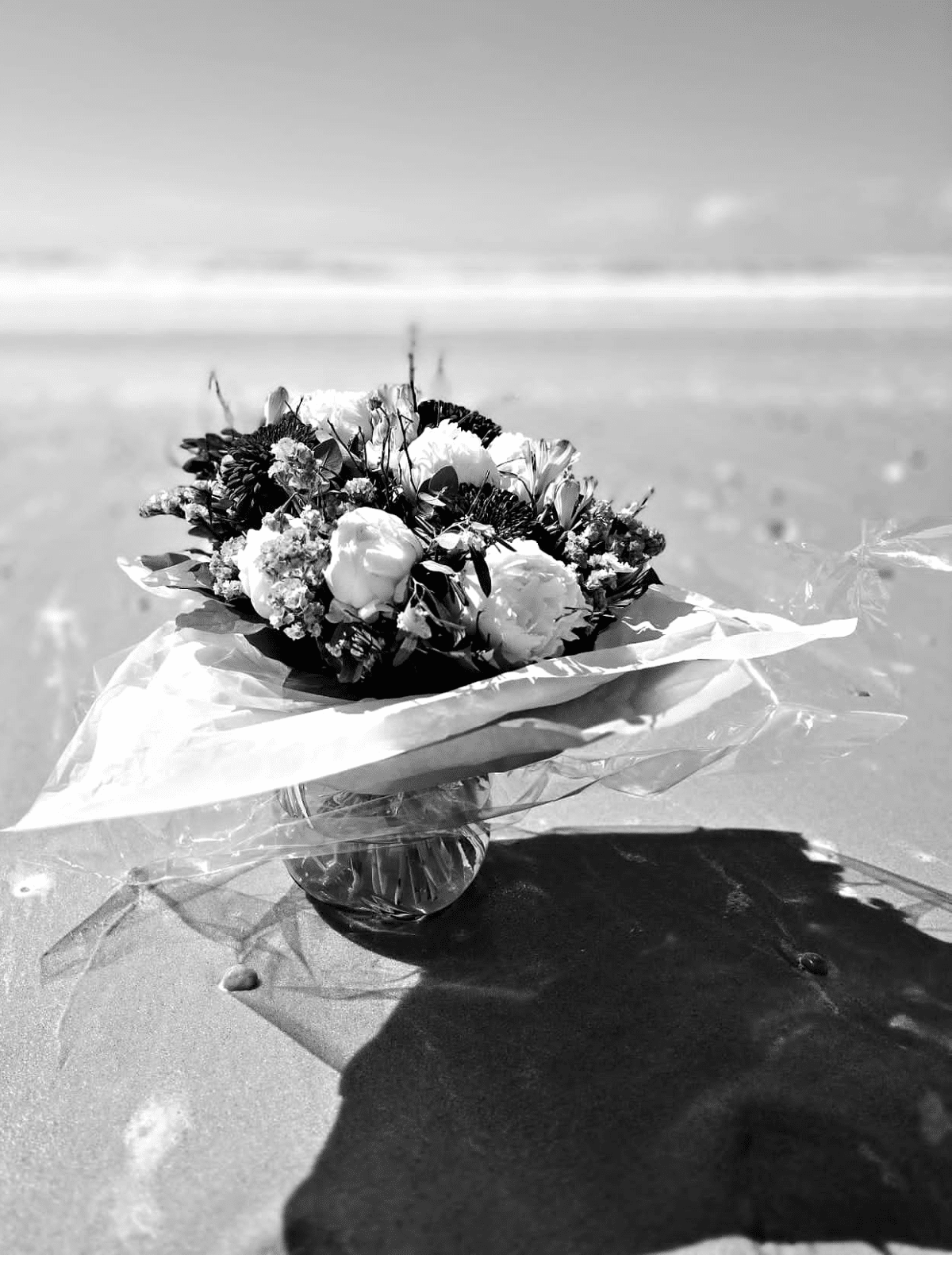 Écho d'Écume_improve Sur le rivage immobile, un bouquet murmure la mer : pivoine en voile d'ivoire et gypsophile pareille à une pluie d'écume, capturant la lumière comme une mémoire fragile.Le verre retient le souffle salin et le sable peint des ombres — une élégie photographique où texture et silence composent une confidence.